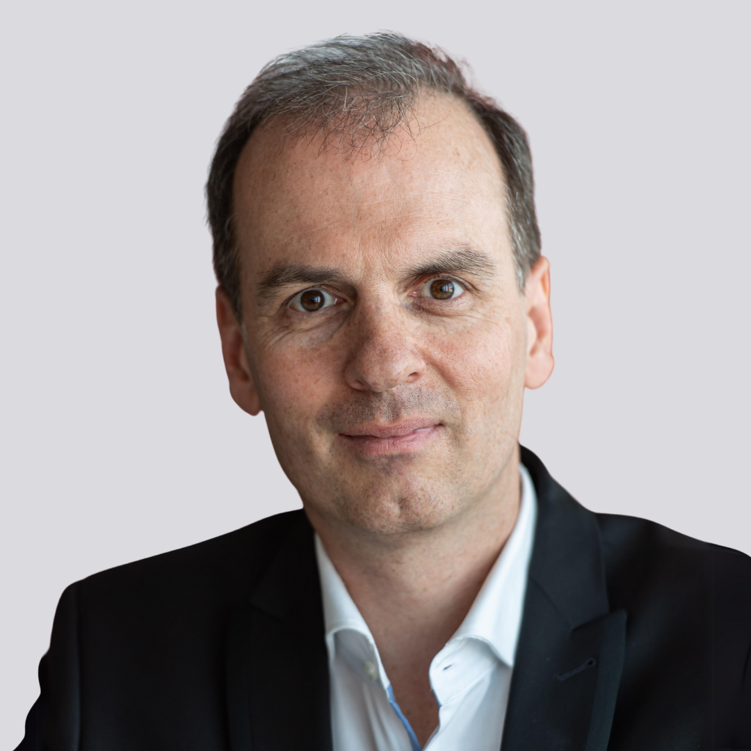 Mann in schwarzer Anzugsjacke und weißem Hemd, der mit neutralem Blick in die Kamera schaut.